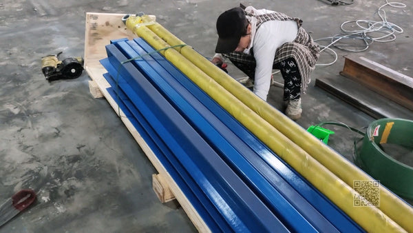 Worker packing blue guide rails and yellow-wrapped lift components on a wooden crate base for shipment